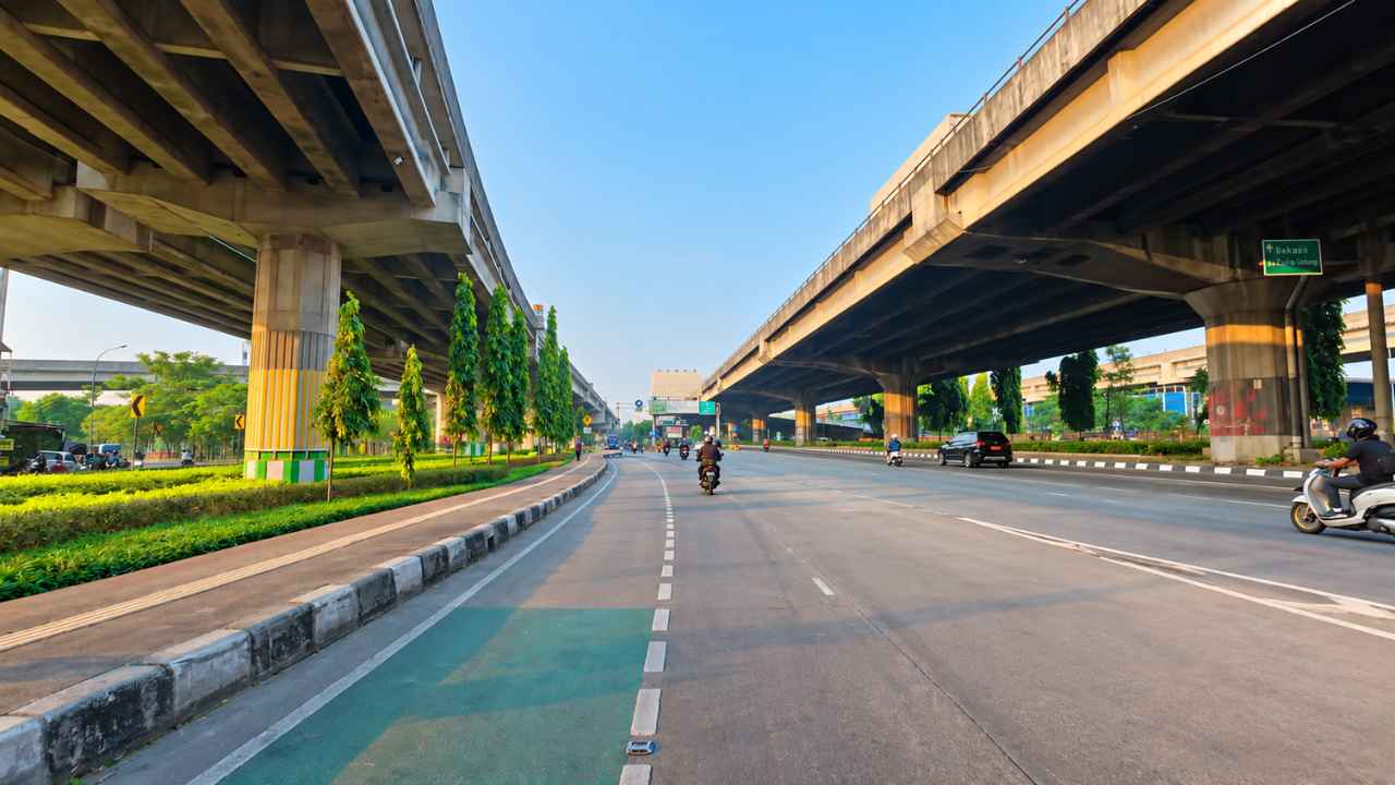 Motorcycles and cars on a broad road beneath elevated concrete spans, with planted verges, roadside trees and blue sky