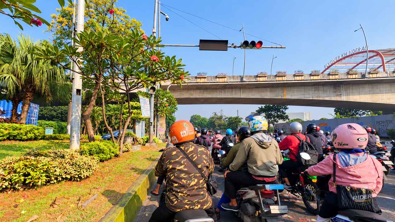Riders on motorbikes waiting at a red light under the concrete span of Flyover KH Noer Ali in Bekasi, with the flyover name board and flowering trees on a clear morning