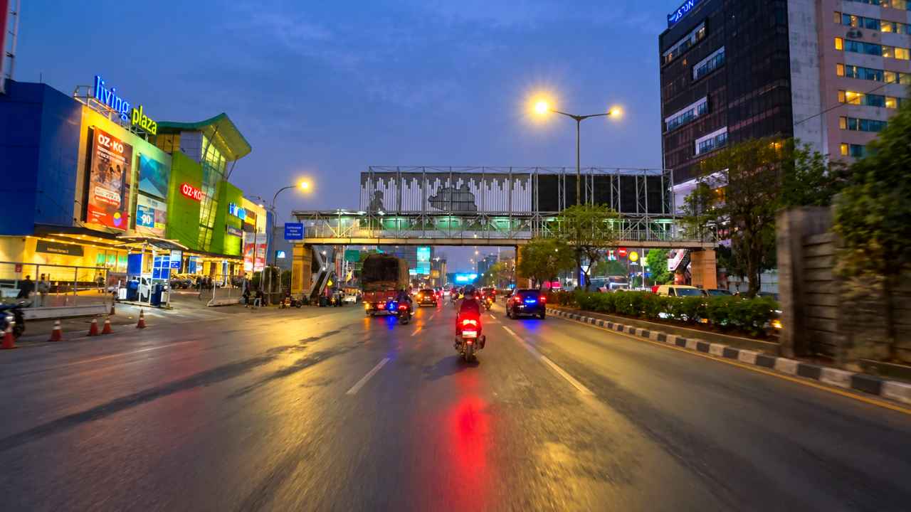 A motorcyclist riding through night traffic on Jalan Ahmad Yani in Bekasi, with the pedestrian bridge (JPO) ahead and the illuminated Living Plaza Bekasi building on the left