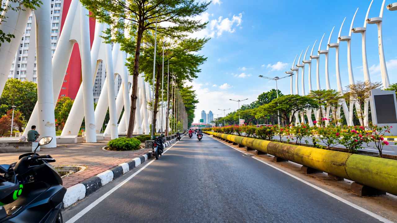 White curved steel pillars of the Kemayoran Gate along both sides of a wide Jakarta road, with motorcyclists, trees and a yellow pipe barrier in the foreground