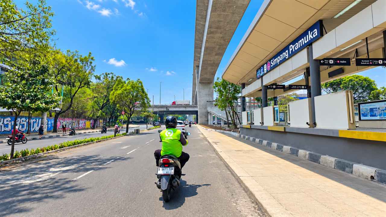 A rider in a green vest on a motorcycle heading along Jalan Pramuka Raya in Jakarta, passing the Simpang Pramuka Transjakarta stop under an elevated structure on a clear morning