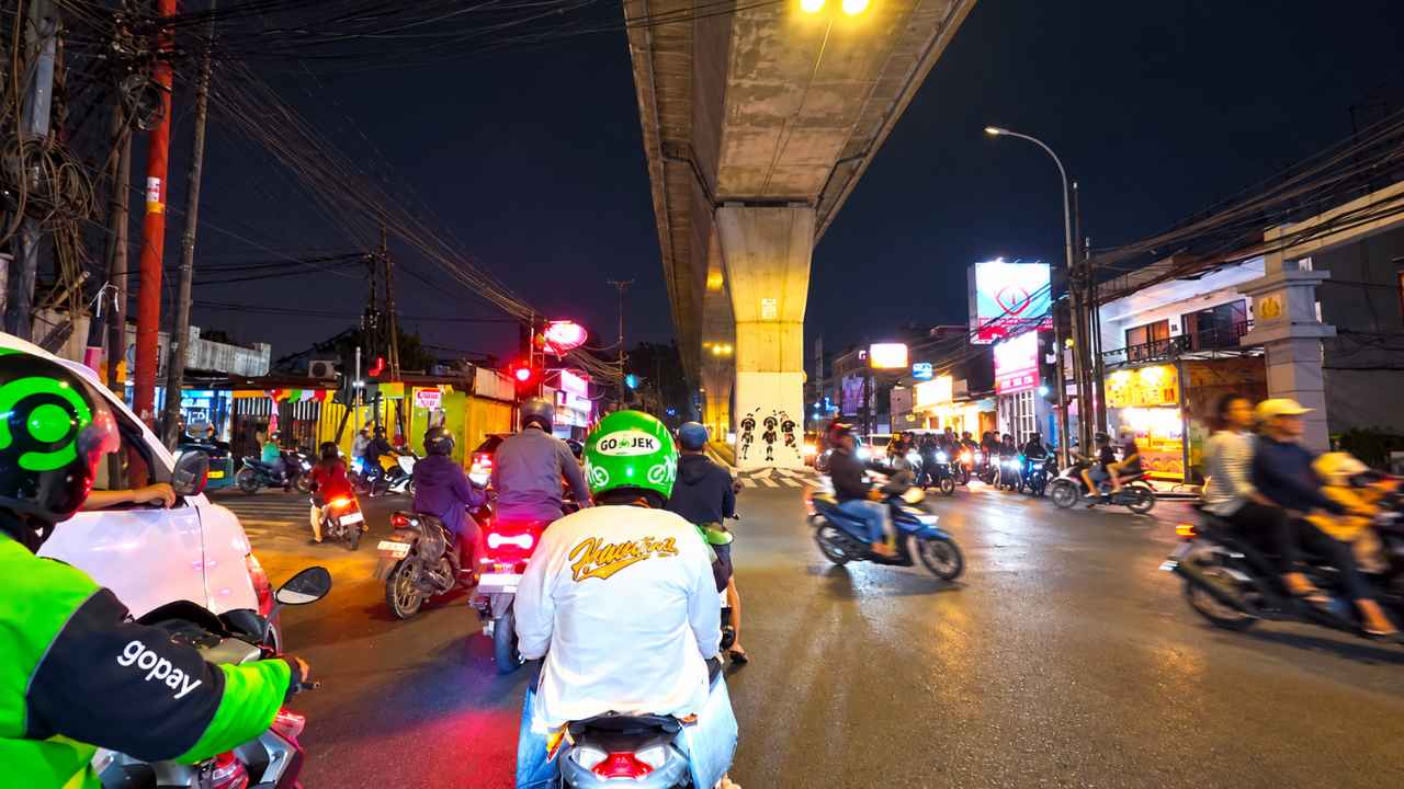 The Seskoal traffic light at night in Jakarta with many motorcyclists, including a ride-hailing driver in a green helmet, under a flyover and lit billboards